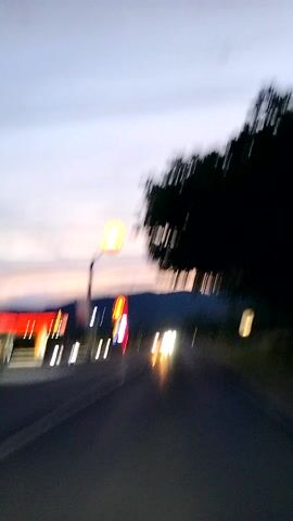 The video shows a view from a vehicle moving along a road at dusk. On the left side of the road, there is a tree and a gas station with illuminated signs. On the right side of the road, other vehicles are visible with their headlights on. The sky is a gradient of blues and oranges, indicating the transition from day to night. The video is shot vertically and is oriented as if the viewer is looking sideways.