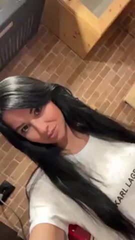 A woman with long, black hair is taking a selfie video in what appears to be a kitchen. She is wearing a white t-shirt with "KARL LAGERFELD" printed on it. She is looking at the camera with a slight smile and runs her hand through her hair at one point. The background shows a brick wall and kitchen appliances.  A grey oven range hood is visible above her and to the left and a wooden cabinet with glass doors is visible above her and to the right.  An electrical outlet and plug can be seen near the bottom left corner of the frame.