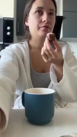 A fair-skinned woman with brown hair is sitting at a table in a kitchen. She's wearing a white robe over a gray tank top. She's eating a piece of chocolate and drinking from a blue ribbed mug. The kitchen has light-colored countertops and white cabinets. There's a black oven in the background. The woman eats the chocolate in small bites, alternating with sips from her mug.