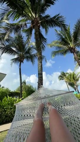 A first-person perspective video shows a person lying in a white hammock. Feet are visible, wearing a silver anklet. The hammock is suspended beneath several tall palm trees. Their fronds sway gently against a bright blue sky with scattered white clouds. Lush green foliage and a portion of a white building are visible in the background.