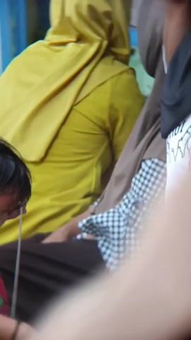 The video shows a child in a red and green shirt with a white string. To their right, a woman wearing a yellow headscarf and a dark brown skirt. Behind them, another woman in a grey headscarf and a pink and white floral patterned shirt. Further in the background, a person wearing a blue headscarf and a plaid shirt is visible, holding a green object. Another person is partially visible in the background wearing a red garment with a black and white patterned scarf. The environment appears to be outdoors, with foliage in the foreground.