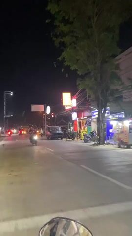The video depicts a nighttime street scene, likely in Southeast Asia, with various vehicles and pedestrians present. The perspective is from a motorcycle, indicated by the visible rearview mirror in the foreground. The street is illuminated by streetlights, neon signs, and headlights from passing vehicles. Several motorcycles are visible, some moving and some parked. A dark-colored van or minivan is seen in the middle distance. There are also a few pedestrians on the sidewalk and crossing the street. The environment appears to be a commercial area, with shops and businesses lining the street. The overall atmosphere is busy and dynamic, characteristic of a lively urban night.