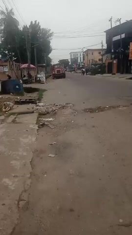 The video shows a street scene with buildings on either side. There are a few people walking on the street, and a red tractor is parked in the distance. There is also some debris on the side of the road. The sky is overcast.