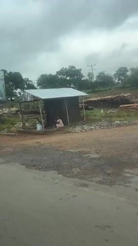 The video pans across a roadside scene in what appears to be a rural or semi-rural area. In the foreground, a paved road shows signs of wear with patches of dirt and debris. To the right of the road is a simple wooden structure, possibly a market stall or small shop, with a corrugated metal roof. Inside and around this structure, several people are visible. One person, wearing a pink shirt and a headscarf, is seated near the entrance. Another person in a grey hooded garment is seated further inside. There are piles of produce, possibly fruits or vegetables, displayed in and around the stall. To the left of the road, a large, damaged billboard stands, displaying political campaign imagery. A person in an orange t-shirt and headscarf walks into frame and gestures outwards. Further down the road, a blue-roofed building and a motorcycle are visible. The sky is overcast, and the surrounding area is covered in vegetation, including trees. Piles of what appear to be lumber or wood are stacked in the background. The overall impression is a snapshot of daily life at a roadside location, possibly a local market or trading post, with elements of commercial activity and political advertisement present.