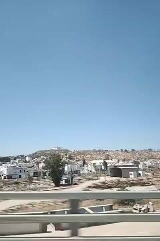 The video shows a view from a moving vehicle looking out at a town or village. The town is characterized by low-rise white buildings, many with red tile roofs. The buildings are clustered together on a hillside, with a larger structure or monument visible at the top of the hill. In the foreground, there is a bridge or overpass with metal railings, suggesting the vehicle is on a road or railway. The landscape is dry and arid, with sparse vegetation. The sky is clear and blue, indicating a sunny day. The overall impression is of a rural or semi-urban settlement in a dry climate.