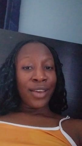 A woman is looking into the camera, smiling, and singing “Thank You Jesus. Thank you my Lord.” The woman has dark hair in braids and is wearing a yellow tank top. The background consists of a dark wood-like surface and a light-colored wall. The video is taken from a slightly angled, close-up perspective.