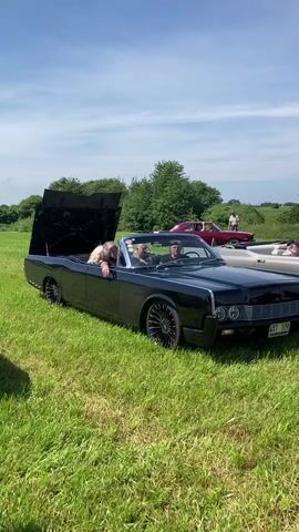 A black convertible Lincoln Continental is parked on a grassy field under a bright blue sky with scattered clouds. The trunk is open, and a person with blonde hair is leaning out of the driver's side, looking back. Two other people are visible in the front seats of the car. In the background, there are trees and other parked vehicles, including a red car and a white boat. The grass is green and appears to be recently mowed. The overall scene suggests an outdoor gathering or car show on a sunny day.