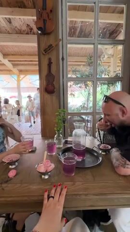 The video shows a group of people sitting around a wooden table, possibly in an outdoor cafe or restaurant. In the foreground, a person's hand with red nail polish rests on the table. To their left, a young girl with blonde hair is holding a pink drink and looking towards the left of the frame. In front of her are two small bowls of pink ice cream. Across from her, a man with a shaved head and tattoos on his arm is leaning over the table, eating something from a small bowl. Between the two people, there are two glasses of pink liquid and a jug filled with the same liquid, all placed on a metal tray. The table is made of wood and has a textured surface. Behind the table, there is a wooden pillar decorated with two violins, one large and one small. To the right of the pillar, there is a large window with multiple panes, offering a view of greenery outside. In the background, through an opening, several people can be seen in a lighter, more open area. One person, a woman with red hair, is walking away from the camera while holding a tray. Another person, a young boy, is walking towards the camera. The overall atmosphere seems relaxed and casual.