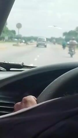 The video shows a person's hands on a steering wheel, with a view through the windshield of a road. A white SUV is driving ahead on the road, with several motorcycles following behind. The road is paved and has lane markings. There are trees and buildings visible on the sides of the road. The sky is cloudy.