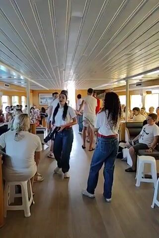 The video shows an indoor scene that appears to be the interior of a boat or ferry. The ceiling is made of white, parallel panels with recessed lighting. The walls are a light tan color, and a wooden floor runs the length of the space. In the background, there are windows with a view of a sunny outdoor scene, possibly islands or coastal scenery.

Several people are visible in the video, most of whom are wearing white t-shirts and blue jeans. Some are gathered in small groups, while others are standing in a line. There's a sense of activity, with some individuals appearing to be interacting, possibly dancing or participating in a group activity. One woman in the foreground, wearing a white t-shirt and blue jeans, has a red sash tied around her waist. Another woman in the foreground, also in a white t-shirt and blue jeans, has curly dark hair and a red sash tied around her waist. A man in the background, wearing a white t-shirt and blue jeans, is looking towards the camera. The overall atmosphere seems lively and social.