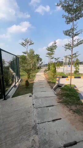 The video shows a paved walkway running alongside a grassy area with several trees. A black chain-link fence is visible on the left side of the walkway. Beyond the grass and trees on the right is a paved area that appears to be a parking lot, with yellow and black parking stall markers and some streetlights. The sky is blue with scattered clouds. The camera is moving forward along the walkway.