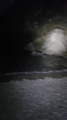 The video shows a very dark beach at night. A flashlight illuminates the shoreline, revealing a rocky beach and water lapping at the shore. There are large rocks visible in the water. The sky is dark and featureless. The camera moves slightly, showing more of the shoreline and rocks. The overall scene is very dark and the details are difficult to discern.