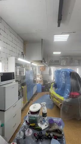 The video shows the interior of a workshop or garage. A car is covered with a blue tarp and yellow masking tape, suggesting it is being prepped for painting or has just been painted. Various tools and supplies are visible on a table in the foreground, including spray bottles, sanding pads, and what appears to be painting equipment. Shelving units with items are present along the walls, and fluorescent lighting illuminates the space. The overall impression is of a working environment dedicated to automotive detailing or repair.