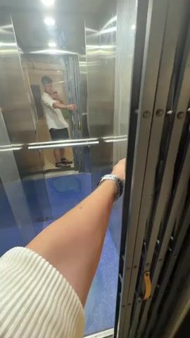 A person with a watch on their left wrist is closing the accordion gate of an elevator. The elevator has mirrored walls and a blue tiled floor. The person is wearing a white t-shirt and black shorts. In the reflection of the mirrored wall, another person is visible. The elevator doors open to reveal a brightly lit hallway with tiled floors and a railing.