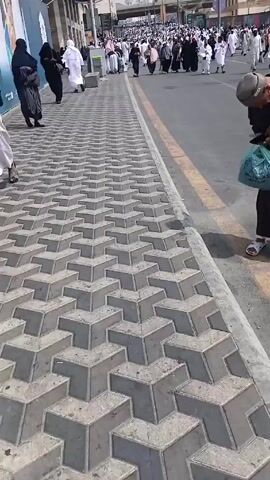 The video shows a street in what appears to be a city in Saudi Arabia with many people dressed in white clothing walking along the street. The people are walking in the same direction. There are tall buildings on either side of the street. The sidewalk has a grey geometric pattern.