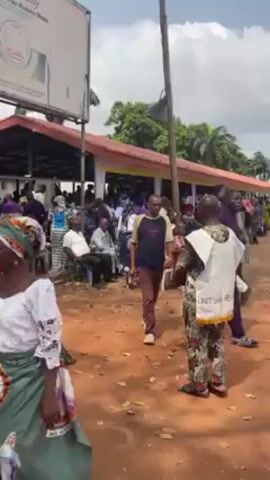 The video captures an outdoor event, possibly a gathering or celebration, taking place on a reddish-brown dirt ground. The scene is filled with people of different ages, dressed in a variety of clothing styles and colors, including traditional attire. Many individuals are walking around, some interacting with each other. A man in a white vest with the words "UNIT SHEL" is standing in the foreground. Another man in a two-toned shirt and brown pants is seen next to him. A person is carrying a large, woven basket with a red cloth inside. A woman wearing a red, black, and gold patterned top and black shorts walks in front of the camera. In the background, there is a structure with a red roof and white trim, possibly a covered seating area. The atmosphere appears lively and social, with people moving about. A sign is visible on the left, although its content is unclear. The overall environment is outdoors, with trees visible in the distance, suggesting a rural or semi-rural location.
