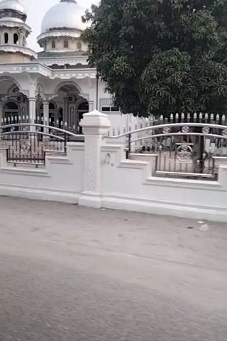 A video shows a white building with a dome and minaret, possibly a mosque, from a slightly elevated perspective. The camera pans to the right, revealing a concrete walkway or courtyard, a white railing with ornate metalwork, and a set of stairs. Further to the right, a smaller, older building with a corrugated roof is visible, along with some vegetation and trees, including palm trees. The overall environment appears to be outdoors, possibly in a tropical or subtropical region, with overcast skies. There is a single person in the foreground near the white building.