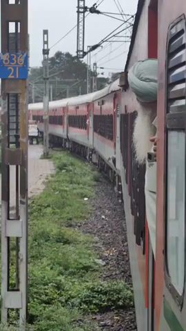 The video shows a train moving on tracks. The train cars are red and white. A man wearing a green turban and a white beard is leaning out of a window of the train. On the left side of the train, there is a pathway made of bricks, and a person is walking on it. There is green grass and bushes on the side of the tracks. Overhead, there are electrical wires and poles supporting them. The sky appears overcast.