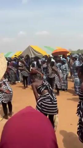 The video shows a crowd of people gathered in an outdoor setting. There are several women wearing black and white striped cloth tied around their waists and black leggings, bending forward and moving to the beat of music. Behind them, there's a larger group of people, some wearing colorful patterned clothing. In the background, several tents or canopies with yellow, green, and white tops are visible. An orange umbrella can also be spotted. The ground appears to be sandy or reddish dirt.