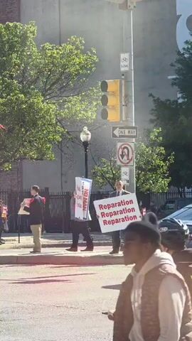A group of people are protesting in the street on a sunny day. Some of them are carrying signs that read "Honk Against Satan" and "Reparation". Others are playing drums in a marching band. Some of the signs say "Satan has NO rights!" and "The Virgin Mary ALWAYS triumphs and crushes Satan's head!". There are trees and buildings in the background. The protesters are standing on the sidewalk next to a busy street with cars passing by.