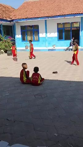 Two children in matching red and yellow uniforms sit on a paved courtyard. In the background, several other children in similar uniforms are engaged in activities. One child is walking, another appears to be playing, and a third stands facing away from the camera. The environment is an outdoor courtyard of a building with a red-tiled roof and blue and white walls. The sun is shining, casting shadows on the ground.