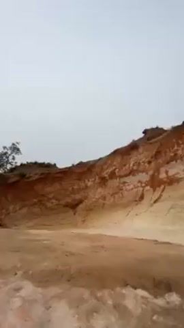 The video pans up along a sand quarry face. The quarry wall has layers of different colored sand. A slender, shirtless man appears in the frame. The man walks on the ground at the bottom of the quarry face.