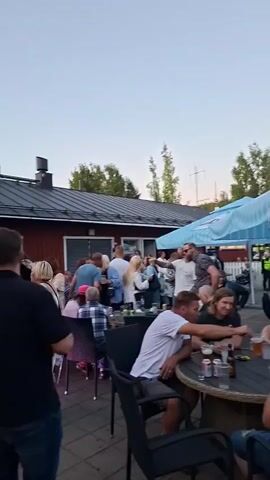 The video shows an outdoor gathering of people near a red building with a black roof. A large sign above the building reads "CAMPING essilä." There are trees and a clear sky in the background. People are standing and mingling, with some appearing to be dancing under pink and green lights. There are tables and chairs set up. A man in a black shirt and jeans is in the foreground, facing away from the camera. Another man in a white t-shirt is seated at a table. Several women are visible, some in dresses and others in more casual attire. The overall impression is of a casual social event.