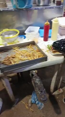 Here is a detailed description of the video:

A street vendor is preparing a food dish on a white table. The table is located outdoors, in what appears to be a public area with other people around. The vendor, wearing black gloves, is assembling the dish on a white plate. The dish consists of french fries, meat, and lettuce.  Bottles of condiments, including ketchup and mayonnaise, are placed on the table. A tray of french fries is also visible. The vendor is adding what appears to be extra meat to the dish. The person is wearing black pants and black shoes with white soles. In the background, other individuals are walking around.