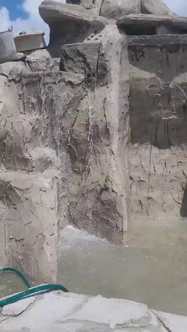 A video shows a partially constructed artificial waterfall. The waterfall structure is made of gray concrete textured to resemble rock. Water is flowing down the rock face and into a pool at the bottom. A green hose is visible in the lower left corner of the frame. In the upper right portion of the video, a person wearing an orange shirt and black pants is visible standing on top of the waterfall structure. They appear to be inspecting the construction. The sky above is partly cloudy.