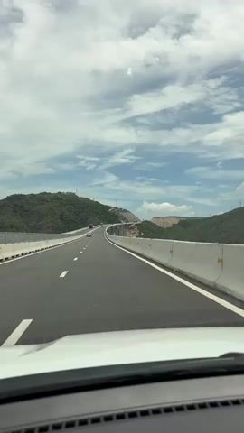 The video shows a first-person perspective from inside a car driving on a highway. The highway is a newly constructed four-lane road with white lane markings. On either side of the highway, there are concrete barriers. Beyond the barriers, there are green, tree-covered hills and mountains. The sky is blue with scattered white clouds. The road appears to be curving to the right and ascending a slight incline. There are a few other vehicles visible in the distance, including what appears to be a red truck and a dark-colored car. The overall environment suggests a scenic, rural location with ongoing infrastructure development.