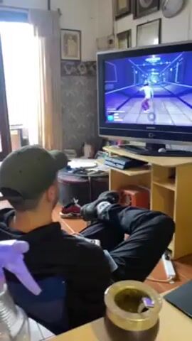 A man wearing a dark green baseball cap, a dark hoodie, and dark pants is sitting in a chair, facing a TV, and playing a video game. The game displayed shows a character walking down a futuristic, blue-lit corridor. The TV is mounted on a light-colored wooden stand, which also has a Playstation console on the shelf below the TV.  To the left of the man is a desk with a spray bottle and a mate gourd on it. Behind the desk and the man is a window with light filtering through the curtains. The room's decor includes framed pictures on the wall and a patterned wallpaper border near the ceiling.  A red and black shoe sits on a circular table behind the man.