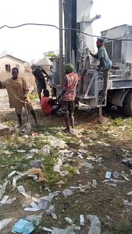 The video shows a group of people working around a large drilling rig mounted on the back of a truck. The rig is positioned near a utility pole, and the area around the truck is littered with trash. Several men are standing around the rig, appearing to be operating or assisting in the drilling process. One man stands on the truck bed, working on the machinery, while others stand on the ground nearby. The surroundings appear to be a residential area, with buildings visible in the background. The ground is covered with grass and trash, creating a cluttered environment. The overall impression is that of a construction or drilling project taking place in a somewhat unkempt location.