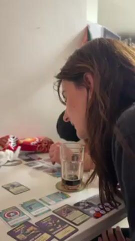 Two people are sitting at a white table, playing a card game. The woman in the foreground is dark-haired, wearing a black hoodie. She has a glass of dark liquid near her. The other person, whose face is mostly obscured, has grey hair and is wearing a black shirt. On the table are several cards, a bag of chips, a coaster, and a crumpled napkin. One of the cards in focus shows a zombie-like figure labeled "YANG".  The people appear to be in a casual, indoor setting.