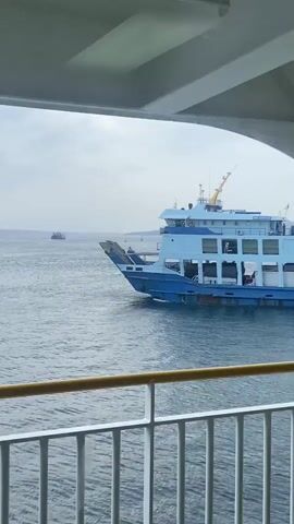 A ferry is visible in the distance on a body of water. The ferry is light blue with white accents and has multiple decks. The front of the ferry has a ramp that is lowered. The water is choppy and appears to be a deep blue. The sky is overcast with a pale grey hue. The foreground shows a white railing with yellow markings. There is also a glimpse of the interior of a vessel, with white ceilings and structural beams.