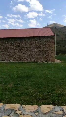The video opens with a view of a stone wall with a red roof. A grassy area is in the foreground. The camera pans to the right, revealing more of the stone building, a large stone wheel, and a cross on the roof. The scene then shifts to a wider view of the area, showing rolling hills, a few trees, and some people walking in the distance. The camera moves again, this time showing a market scene with several white tents, cars, and people browsing. The ground is dusty and there are some green plants and debris scattered around. The sky is blue with scattered clouds.