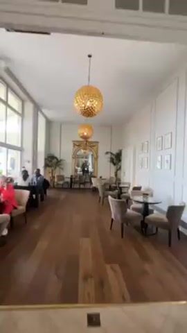 The camera slowly pans across a room in a restaurant. The room is long and narrow, with light brown wooden floors and white walls. There is a large arched mirror with a gold frame on the wall at the end of the room, centered between two doorways. The room is decorated with small round black tables surrounded by tan chairs. Potted plants are placed next to the mirror. There are two large golden globe-shaped chandeliers hanging from the ceiling. There are two people seated at a table near a window. Additional pictures are on the wall to the right of the mirror.