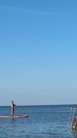 A person is stand-up paddleboarding on calm, blue water under a clear blue sky. They are wearing a dark swimsuit and holding a paddle. The paddleboard is orange. To the right of the frame, there is a section of what appears to be a pool or a contained swimming area with a ladder and stone-like texture. The horizon line is visible in the distance, separating the water and sky.