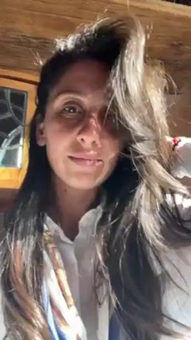 A fair-skinned woman with long dark hair and a white collared shirt is looking directly at the camera. Her hair is partially covering her face. She is indoors, with a wooden beam visible above her. To the left of her, there is a window with a stained glass design. She is holding her hair up, and a red bracelet is visible on her right wrist.