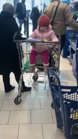 A young child sits in the child-seat of a shopping cart. The child wears a pink jacket, a knit cap, pink pants, and boots. The shopping cart is surrounded by people. The environment appears to be inside of a store with a glass entrance.