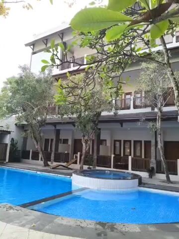 A hotel swimming pool area is shown. The pool is a bright blue and rectangular with a smaller circular spa area attached. A building with multiple levels and balconies is visible behind the pool. Trees and foliage surround the area.