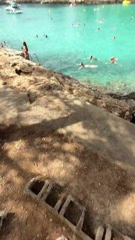 The video shows a crowded beach with many people swimming and relaxing in the turquoise water. The beach is lined with rocky cliffs and trees. In the foreground, there are people sitting on the rocks and looking at their phones. In the background, there are buildings and more trees on the hills overlooking the bay. The water is clear and calm, and the sky is blue and sunny.