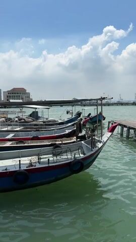The video shows a line of small fishing boats docked at a pier on a sunny day. The boats are of similar size and design, with red and blue paint schemes. They appear to be made of wood or fiberglass, and have small outboard motors attached to the sterns.  The water is calm, with a slight green tint.  In the background, a town can be seen with a tall, distinctive church steeple prominent in the skyline. A building on stilts can be seen to the left of the boats, adjacent to the buildings in the distance. The sky is blue with some white, fluffy clouds. The video has a steady, handheld quality.