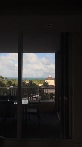 The video shows a view from a balcony, looking out to the ocean. The foreground includes a table and chairs on the balcony, seen through a doorway. Beyond the balcony, there is dense tropical vegetation, a building with a dome, and the ocean. The sky is partly cloudy. There is no visible activity in the video.