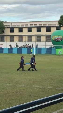 Two medics and a person in a black t-shirt carry an injured person on a stretcher across a grass field. The injured person is wearing a blue t-shirt and blue shorts. The medics are wearing black pants and blue shirts. The person in the black t-shirt is wearing black pants. There is a large green inflatable balloon that says “Apuestas Royal” behind them. There is a soccer goal to the right of the balloon. There is a person in a green t-shirt and green shorts standing in the background to the right. There are buildings and cars in the distance behind the fence. The sky is overcast.