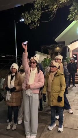 A group of people, some wearing matching beanies and scarves, stand outside a building at night. A woman in the foreground, wearing a pink puffer jacket, grey sweatpants, and sunglasses, holds up a smartphone, appearing to take a selfie or video of the group. Several other individuals, including a man in a yellow puffer jacket and sunglasses, are looking towards the camera. The environment suggests a cold evening, possibly in a town or at an outdoor establishment, with some foliage visible overhead. The building in the background has lights on.