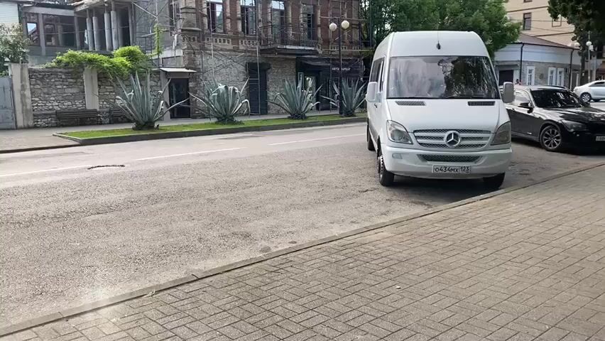 The video captures an exterior street view with a partially renovated building in the background. A white Mercedes-Benz Sprinter van is parked on the right side of the road. In front of the building, there are several large agave plants and a stone fence. Scaffolding surrounds the upper portion of the building, which appears to be undergoing significant renovation. A single person, dressed in a light blue shirt and light-colored shorts, is visible working on the roof. The building itself is a multi-story brick structure with visible architectural details, including ornate window frames and balconies. Two street lamps are positioned in front of the building. A black car is parked behind the white van. The sky is overcast, suggesting cloudy weather. The street is paved, and a sidewalk runs along the right side of the road. The overall impression is a street scene in a town or city, with ongoing construction work on an older building.