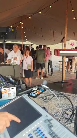 The video begins with a medium shot of a tent with string lights overhead. Inside the tent, people are milling around, some standing and talking, others near a red keyboard and a drum set. The camera then pans to the right, showing the drum set and more of the stage setup. The video then shifts to a wide shot of a sandy beach with the ocean in the background. The sun is setting, casting a warm glow over the scene. The camera pans across the beach, showing sand dunes and some equipment on the ground. The overall impression is of an outdoor event or concert taking place by the sea.