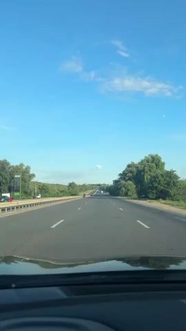 The video shows a first-person perspective of driving on a highway under a clear blue sky with scattered white clouds. The road ahead is paved with white lane markings, and there are trees and greenery on both sides of the highway. In the distance, other vehicles can be seen on the road. The dashboard of the car is partially visible in the foreground. The scene appears to be a rural or suburban highway on a sunny day.