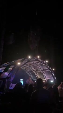 The video shows a nighttime outdoor scene with a DJ performing under a geodesic dome structure. The dome is illuminated with lights that change color, from warm yellow to cool blue. Above the dome, a large, organic-looking object hangs suspended from wires. The object appears to be made of natural materials and has green lights embedded in it. A crowd of people is visible in silhouette, gathered in front of the dome, suggesting a performance or party is taking place. The background consists of trees, indicating the setting is likely a forest or wooded area. The lighting shifts create a dynamic and atmospheric visual effect.