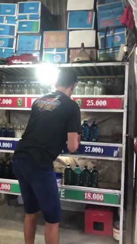 The video shows a man in a black t-shirt and blue shorts standing in front of shelves stocked with bottles and boxes. The shelves are organized with price tags indicating amounts such as 17.000, 25.000, 19.000, and 12.000. The man is reaching for a green bottle on one of the shelves. Behind the shelves, there are more stacked boxes and some corrugated metal roofing. The environment appears to be a retail or storage area, possibly a small shop or a storage unit. The lighting is somewhat dim, with a bright light source illuminating the shelves from above.